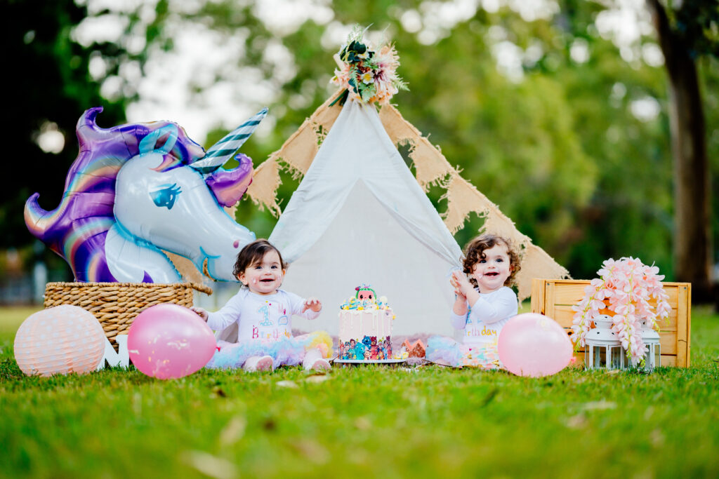 Two little girls joyfully pose in front of a unicorn tent at a cake smash photoshoot